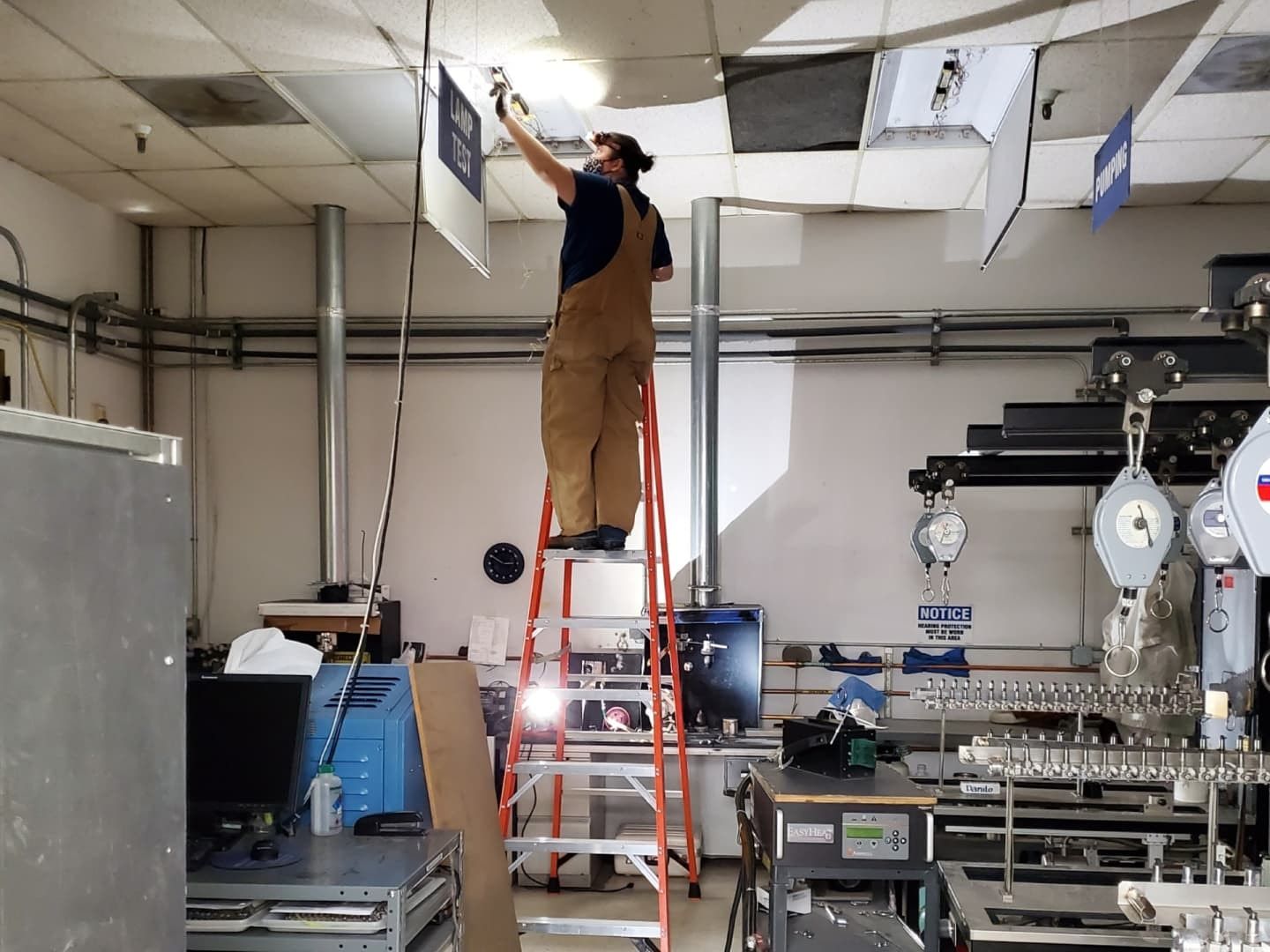 Person on a ladder, installing something on the ceiling in a workshop. Overalls, flashlight, various equipment.