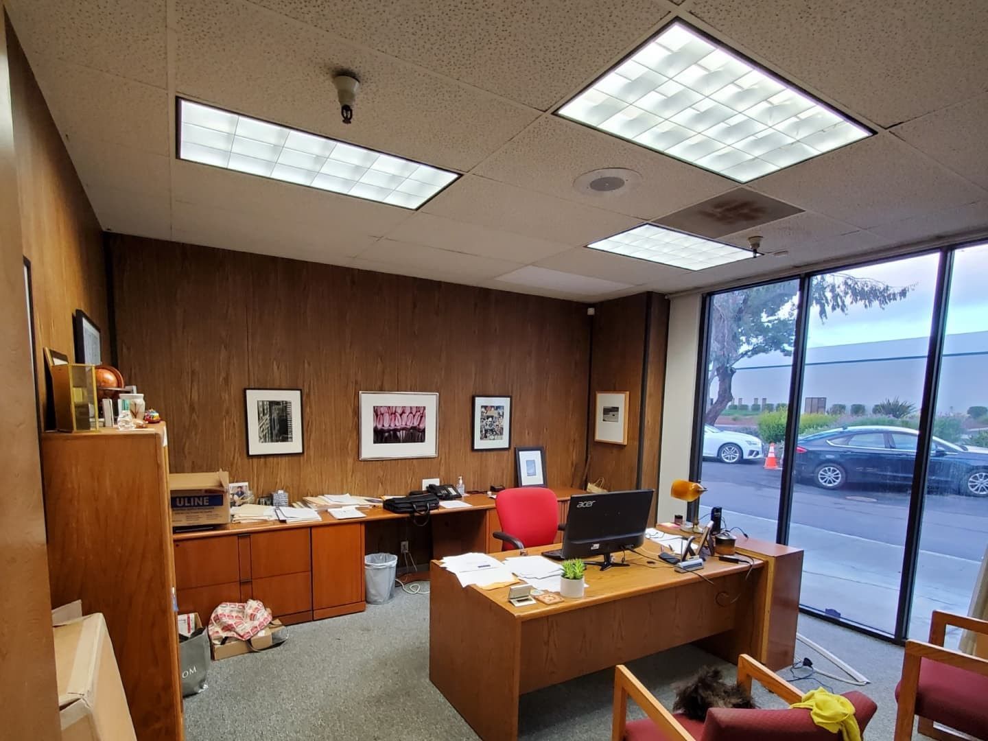 Office with wooden desk, wall, and cabinetry. Windows look out to a parking lot. Red chair, desk clutter, & artwork present.