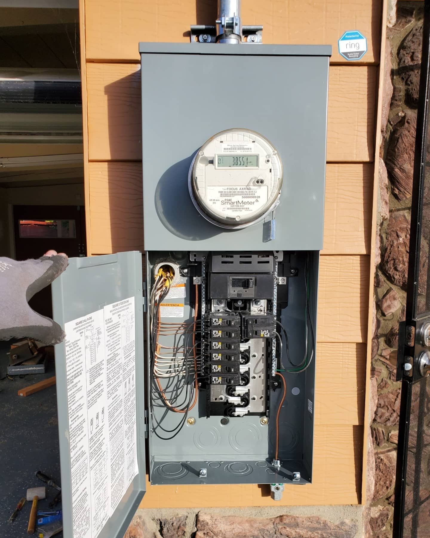 Exterior electrical panel with open door, mounted on a tan-sided building. Wires and breakers are visible.