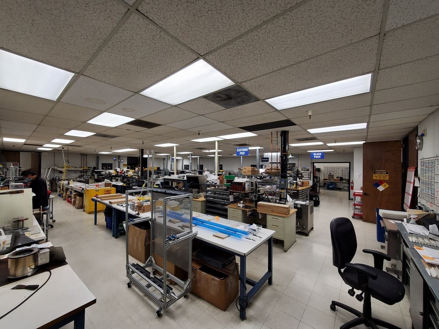 Factory workshop with workstations, equipment, and various supplies under fluorescent lights.
