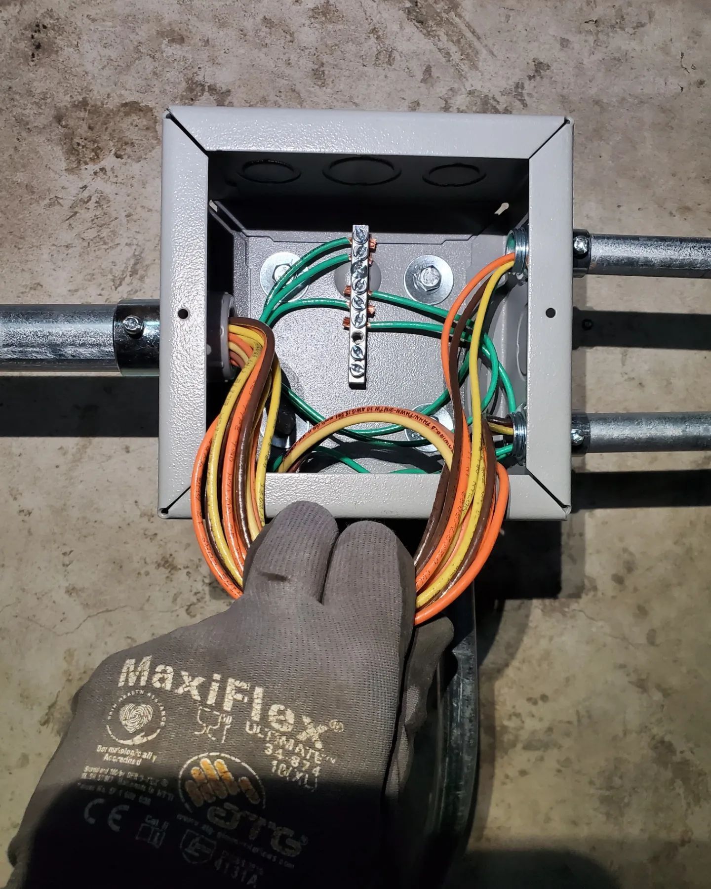 A hand wearing a work glove holds colored wires at an electrical junction box with green ground wires.