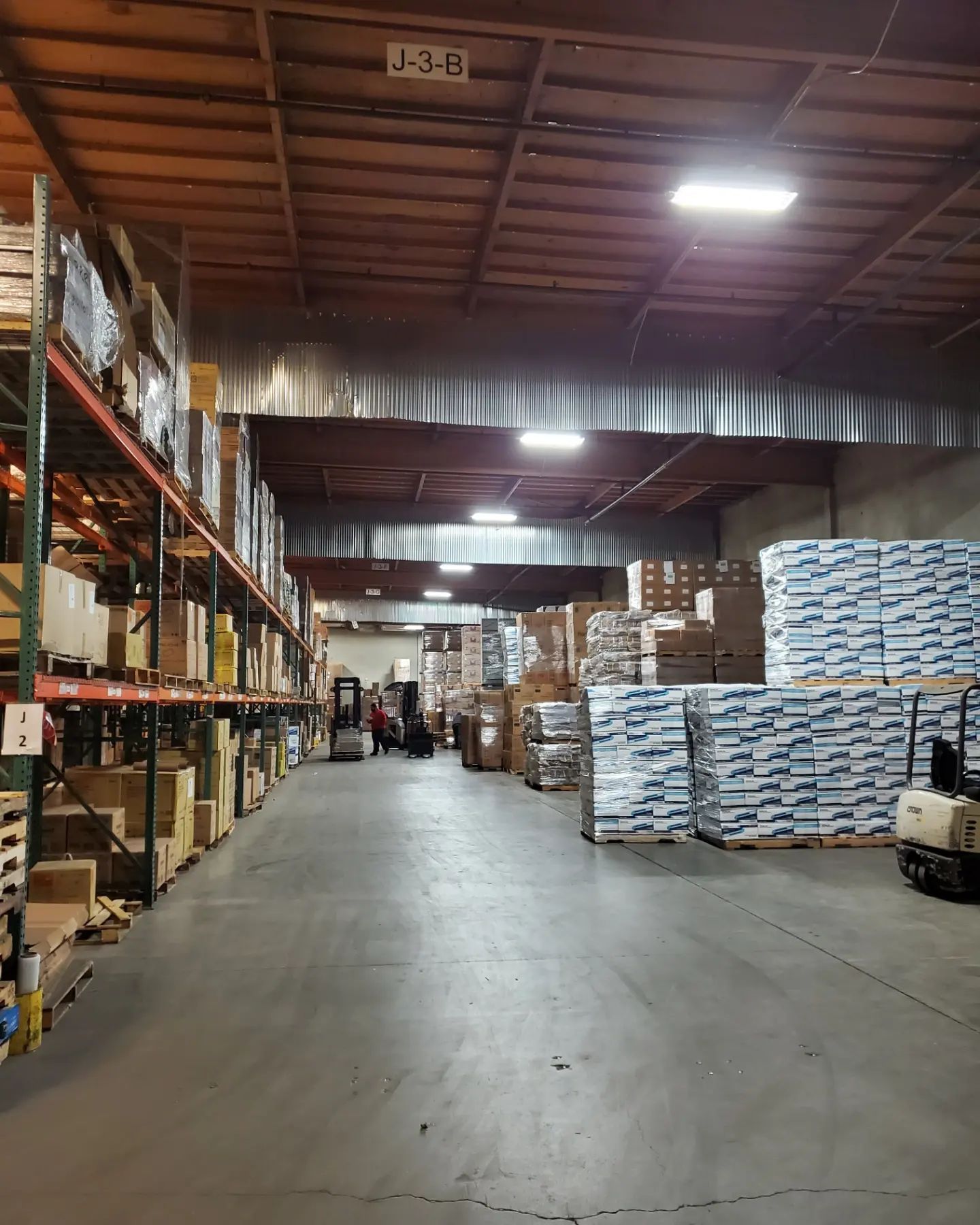 Warehouse interior with tall shelves stocked with boxes and pallets, and workers in the distance.
