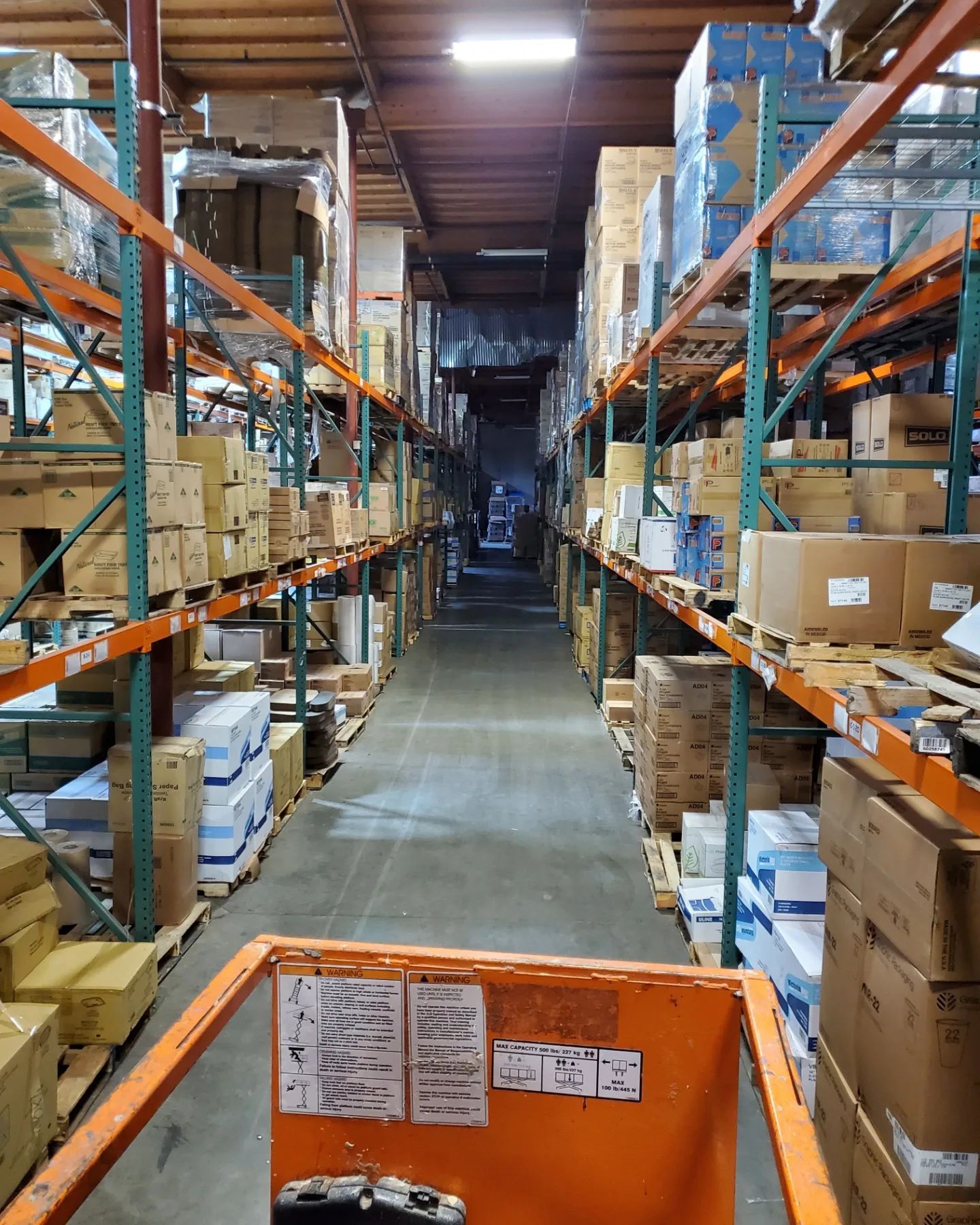 Warehouse aisle filled with shelving units holding boxes; a lift truck is in the foreground.
