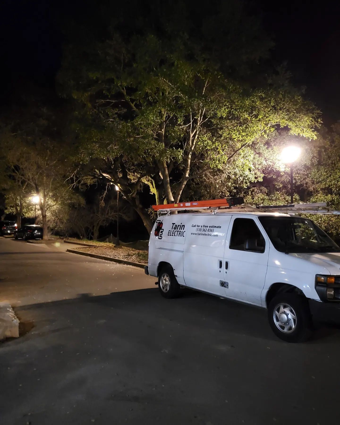 White service van parked at night under a tree, streetlights illuminate a dark area.