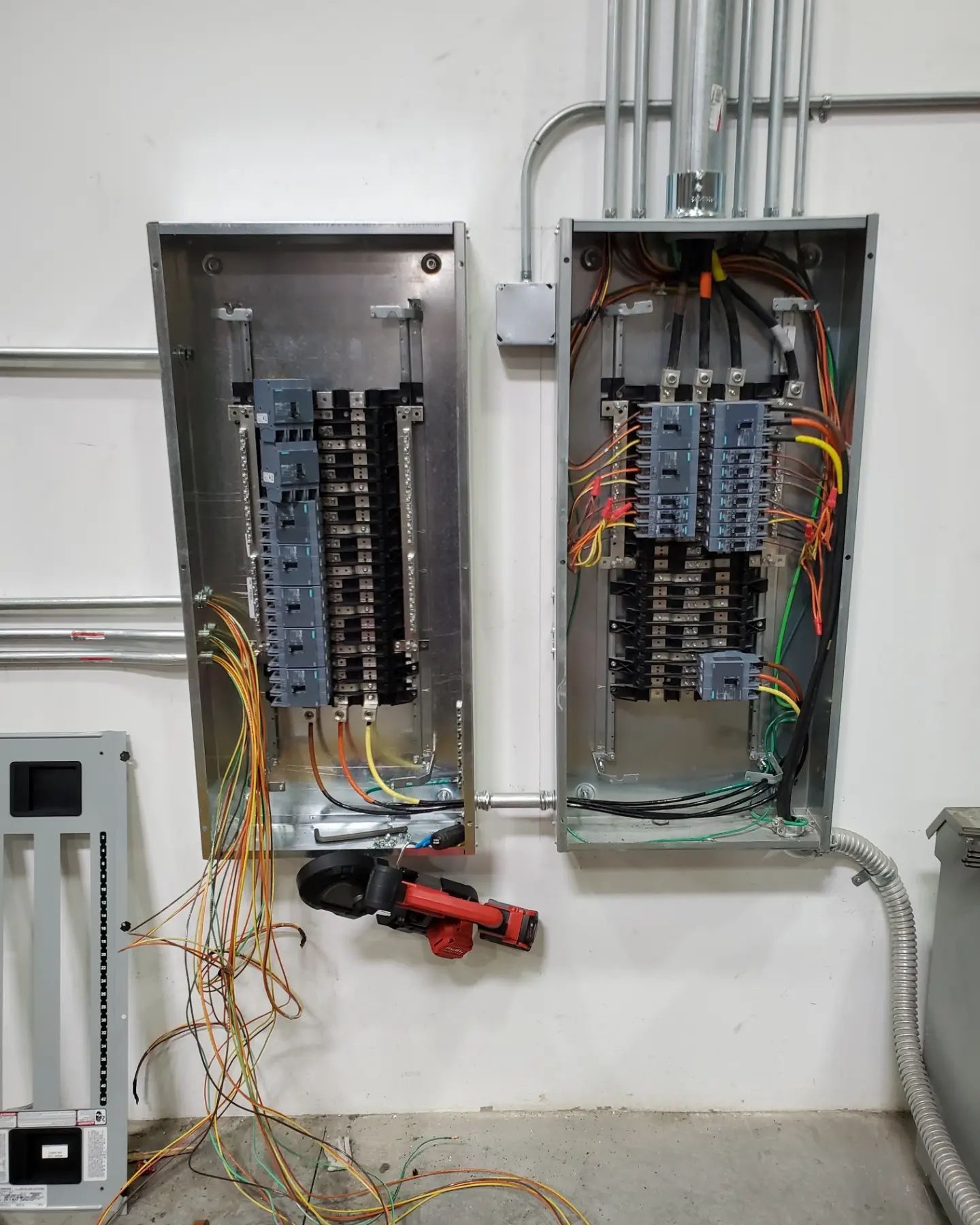 Two open electrical panels with wiring and conduit on a wall. A grinder is on the floor.