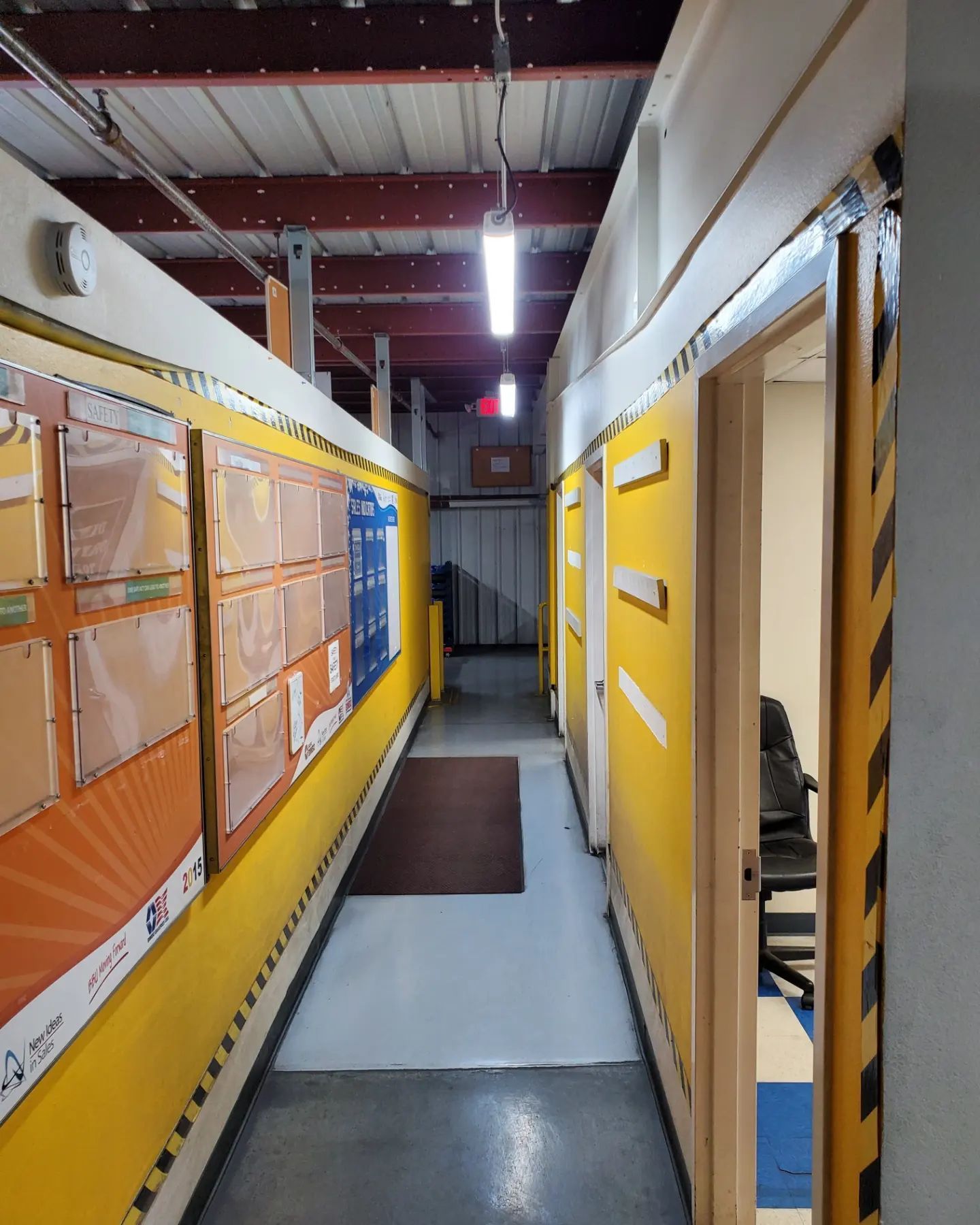 Narrow hallway with yellow walls, doors, and a brown mat. Fluorescent lights illuminate the space.