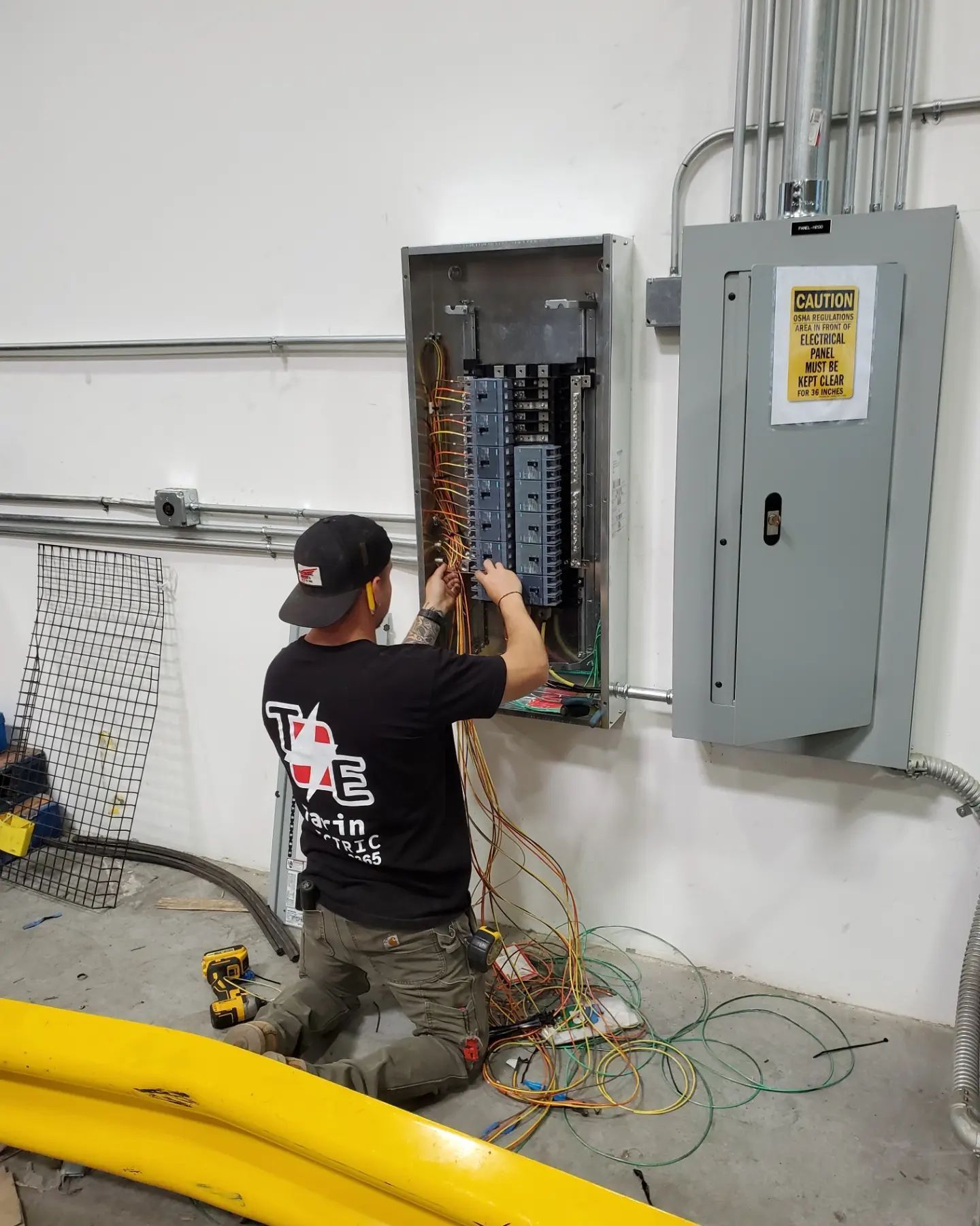Electrician kneeling, wiring an electrical panel on a wall. Wires and conduit are present.