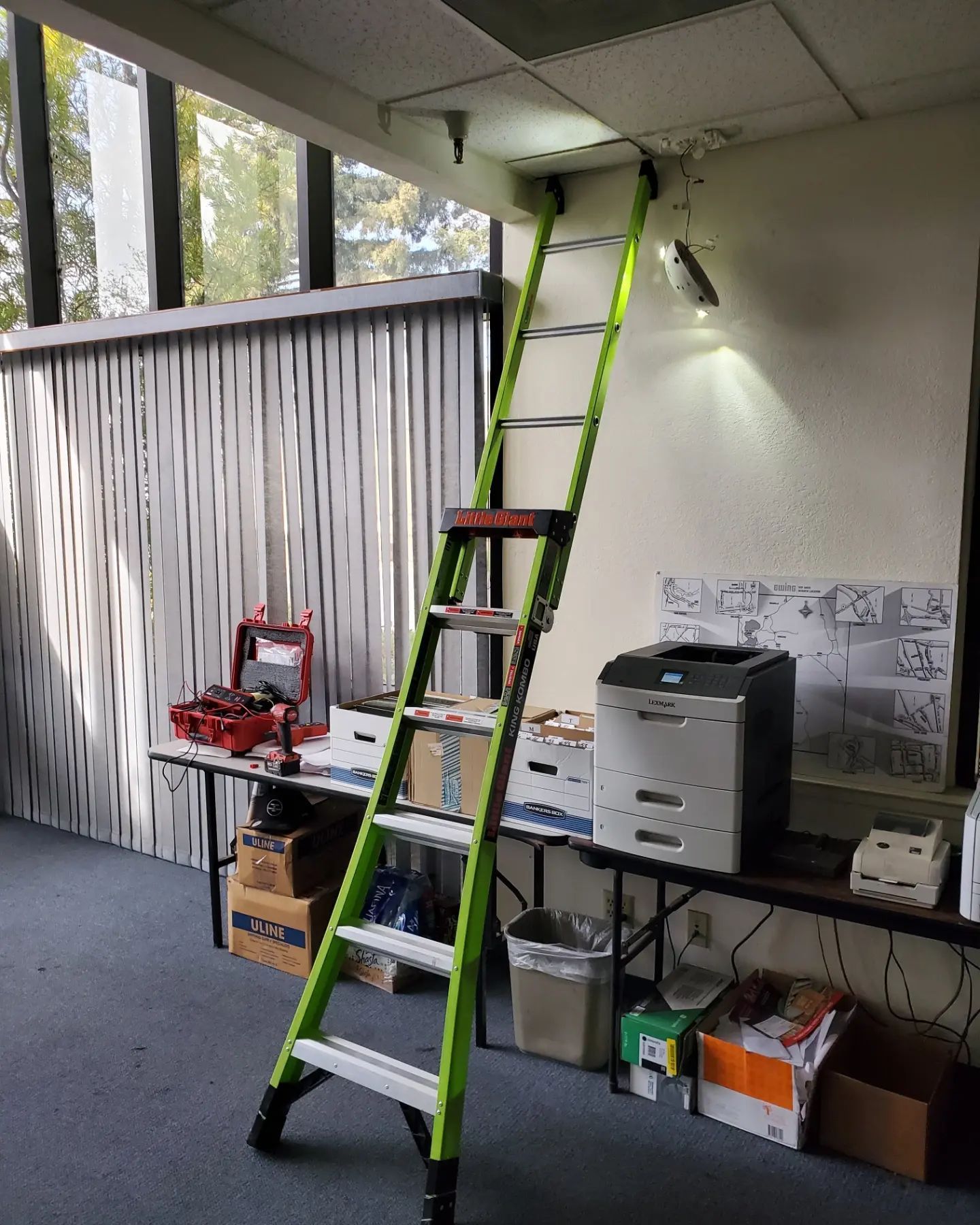 Green ladder leaned against a white wall. Workspace has a printer, tools, and a window.