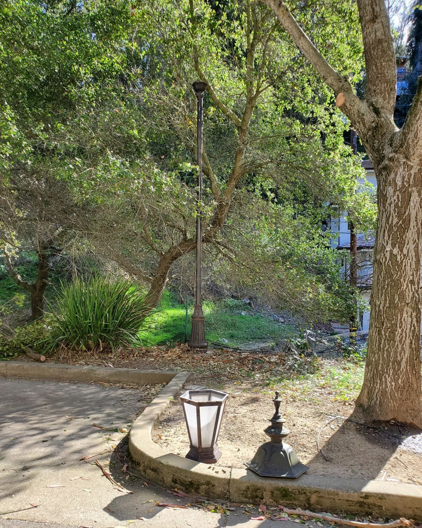 A tall black lamp post with a broken lantern on a cement wall with trees in the background.