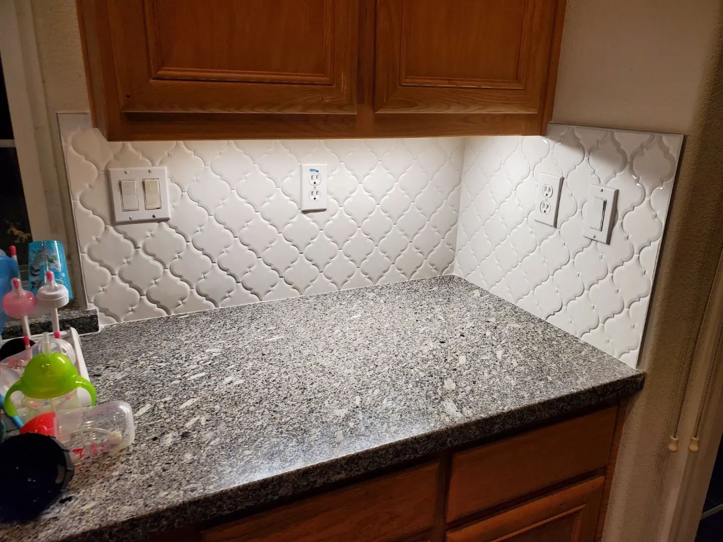 Kitchen backsplash with white patterned tiles, granite countertop, wooden cabinets, and outlets.