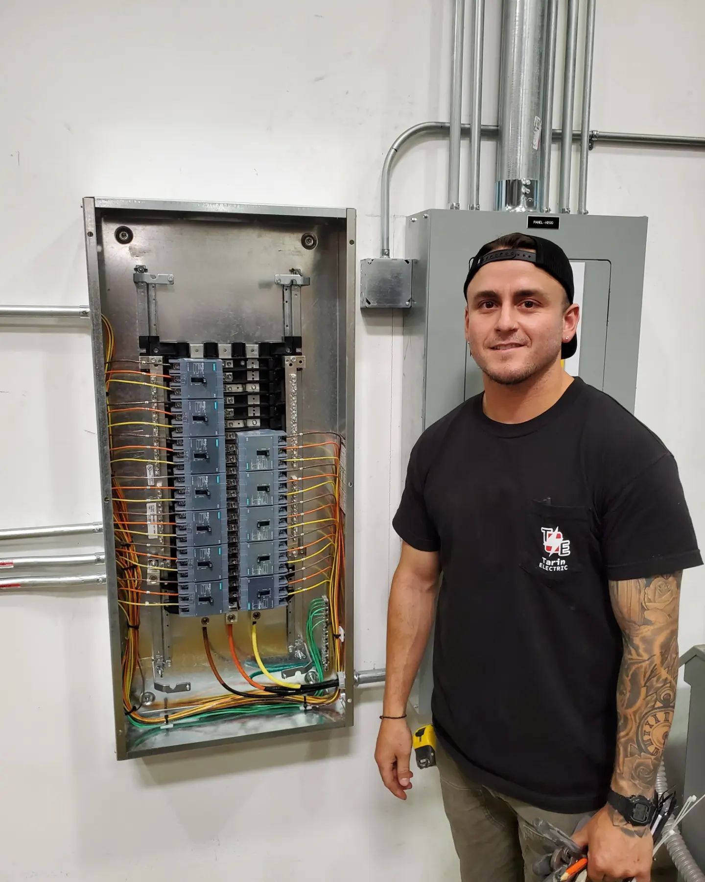 Man stands next to electrical panel. Wires visible.  He wears a black t-shirt, a cap, and holds tools. Industrial setting.
