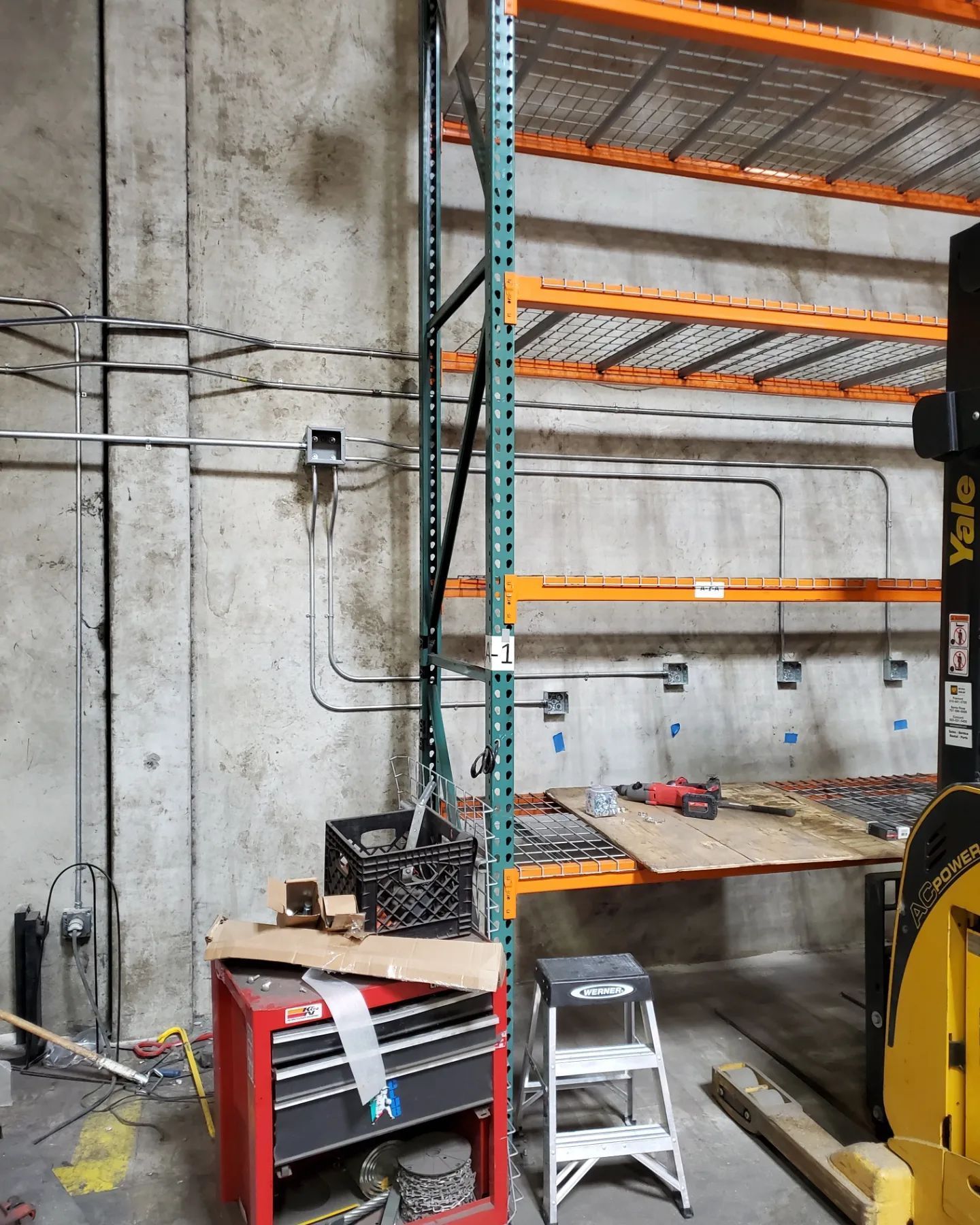 Warehouse interior with shelving, electrical conduits, toolbox, step stool, and forklift.