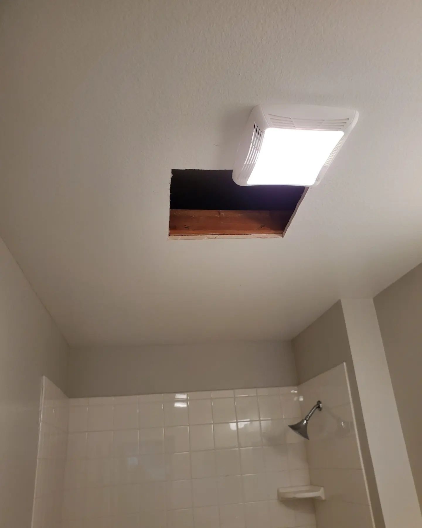 Hole in bathroom ceiling next to a light fixture, above a shower with white tiles.