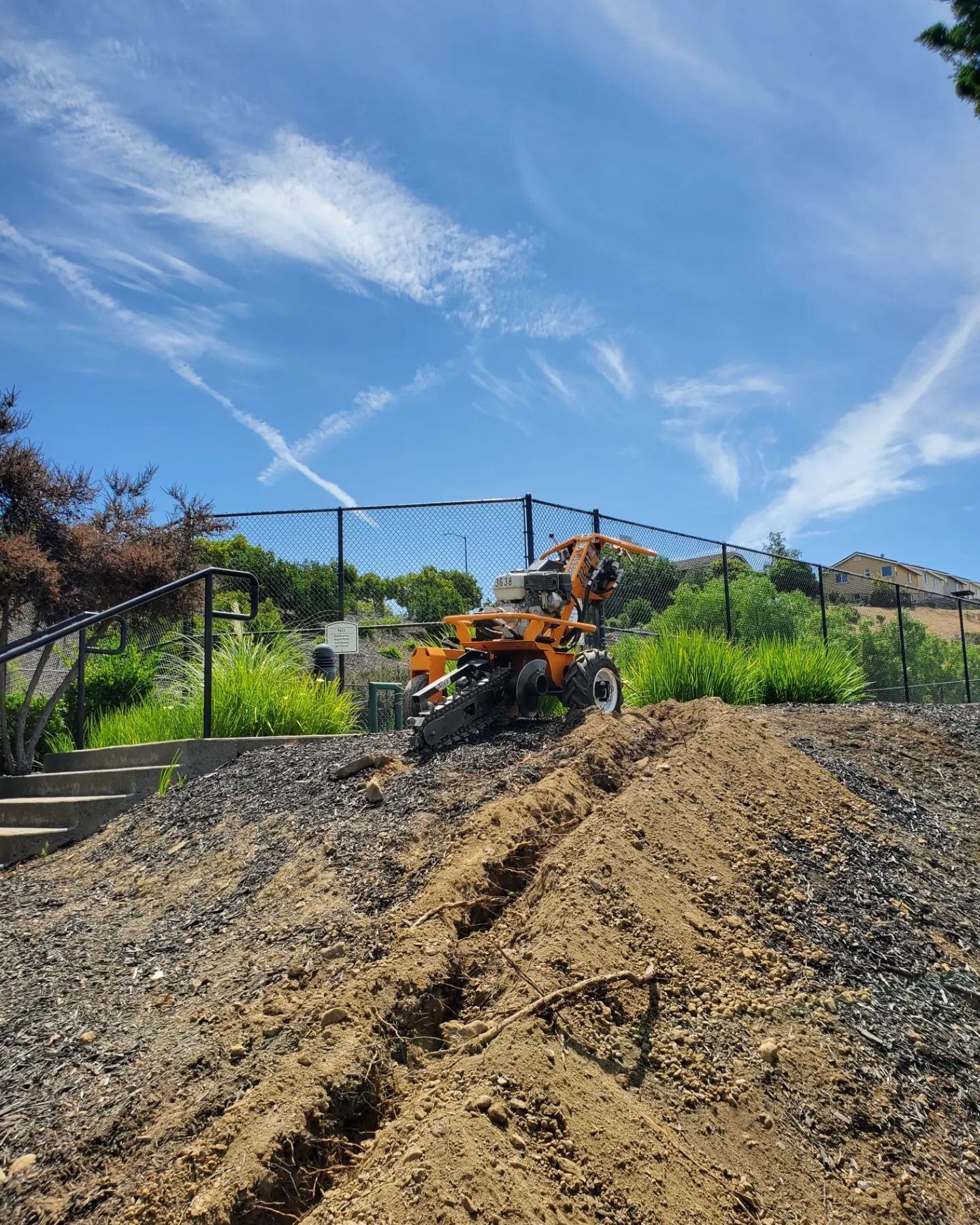An orange tiller digging a trench on a hillside under a blue sky, next to a gravel path.