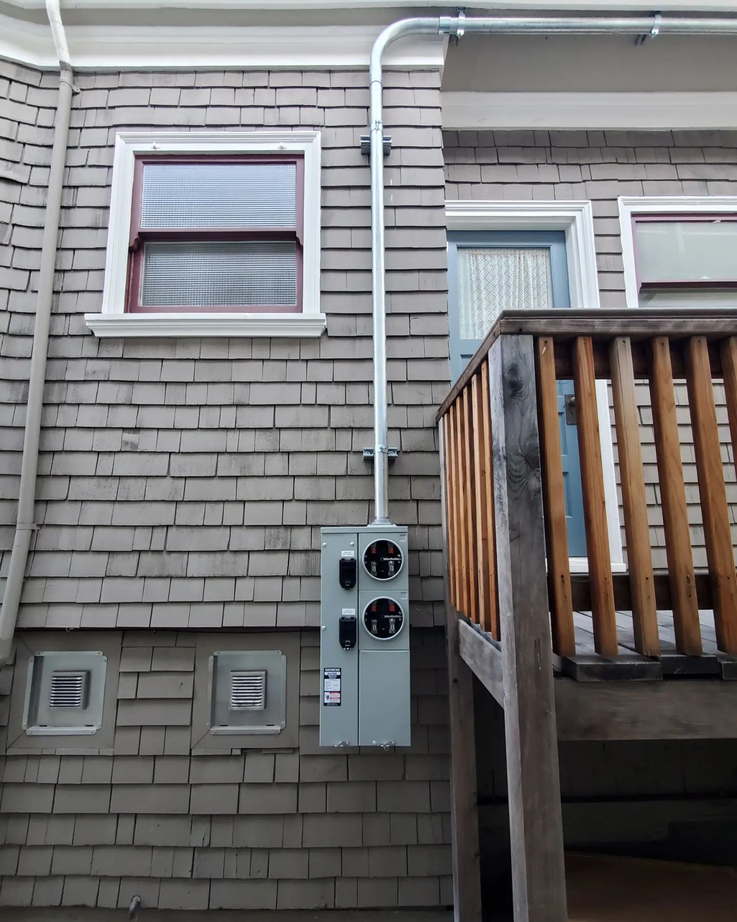 Exterior electrical panel on a shingled wall, with conduit running upwards to the right.