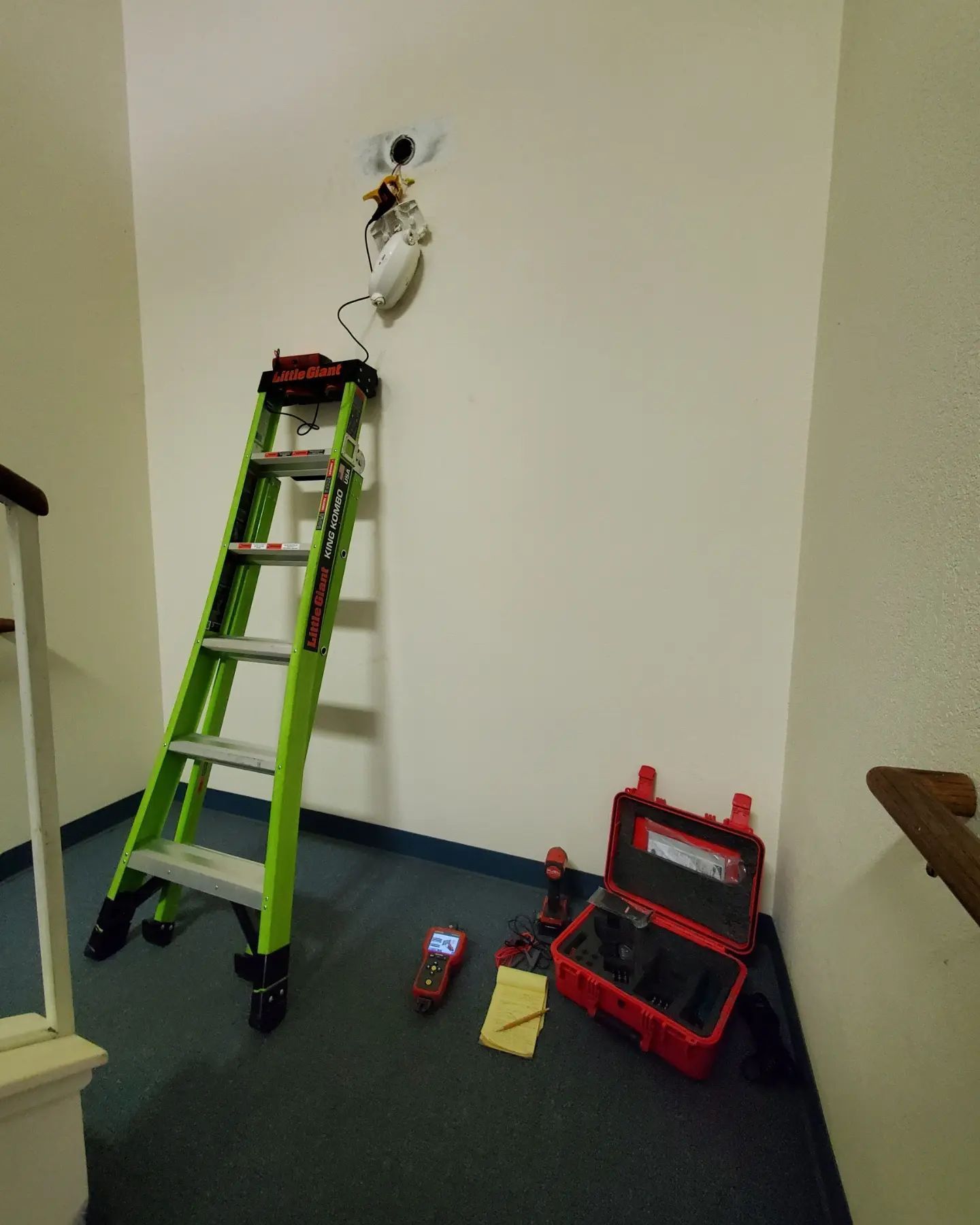 A ladder, tools, and a security camera are set up on a wall in a hallway.