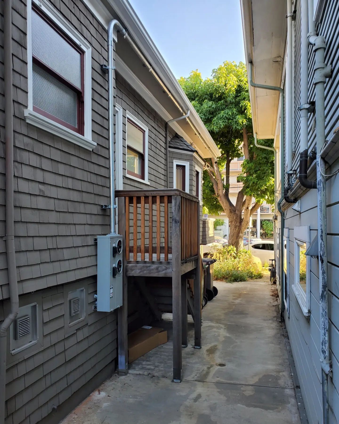 Narrow alleyway between two buildings. One with gray siding and a wooden deck, the other with light blue siding.