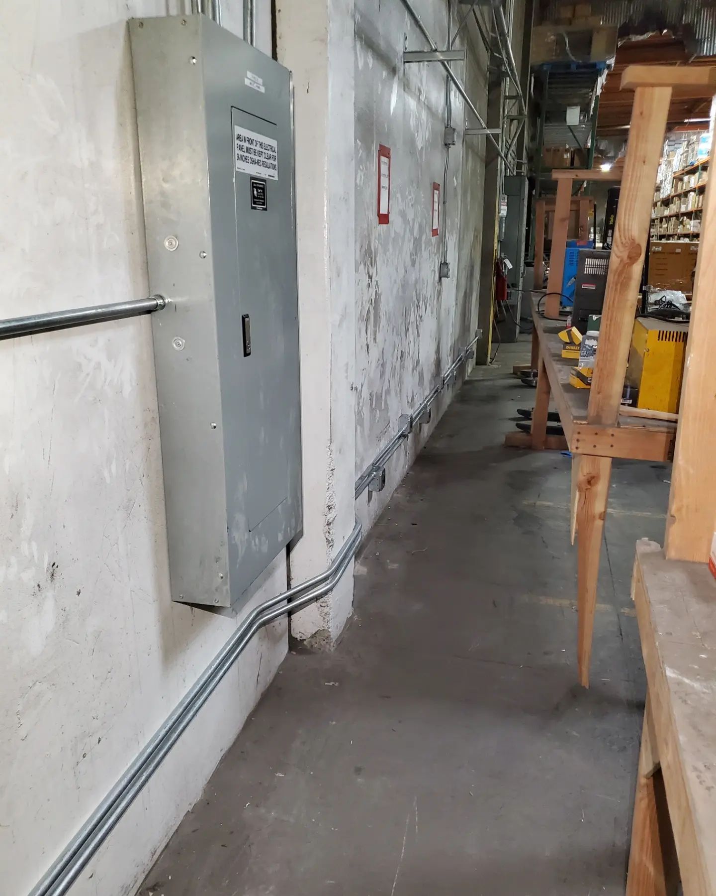 Electrical panel on a concrete wall in a workshop hallway with conduit and workbenches.