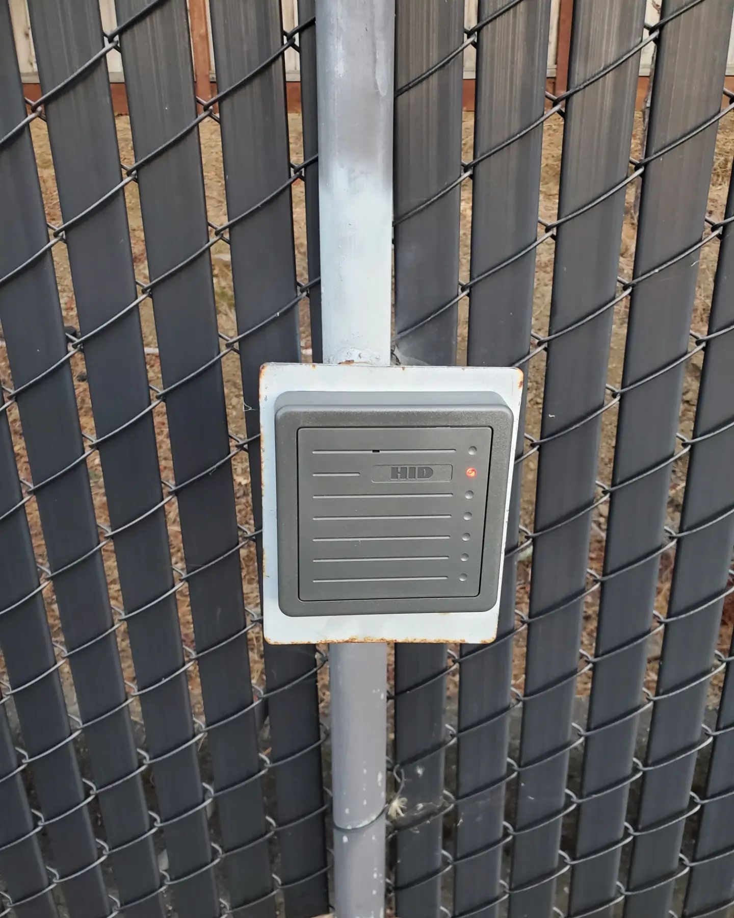 Black RFID access reader mounted on a gray metal post, attached to a black chain-link fence.