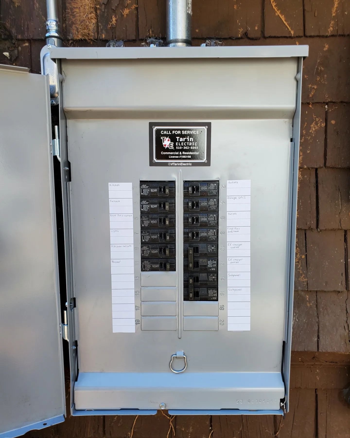 Electrical panel with open door, mounted on a brown wall. Silver metal panel with circuit breakers and labels.