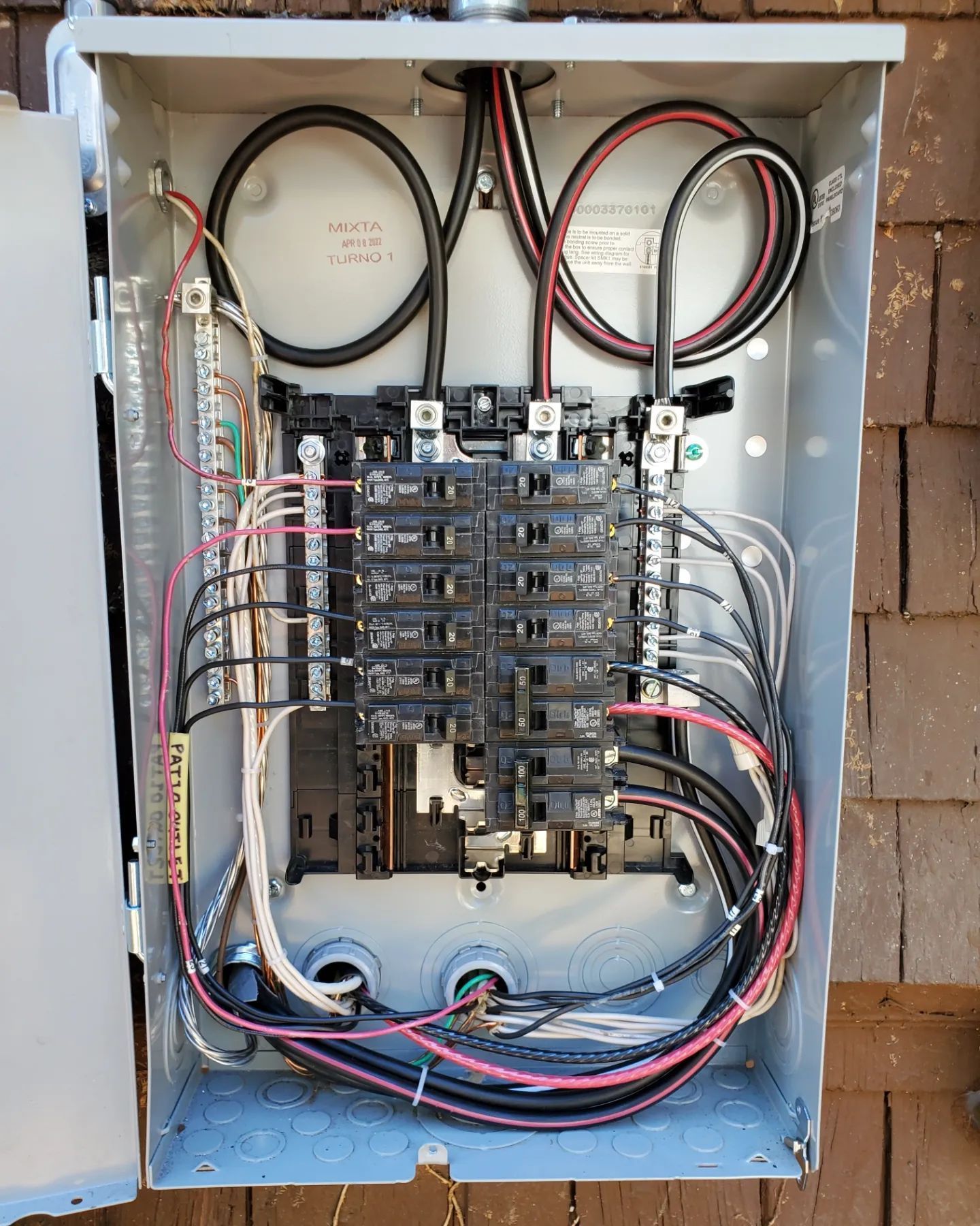 An open gray electrical panel with various colored wires and breakers installed on an exterior wall.
