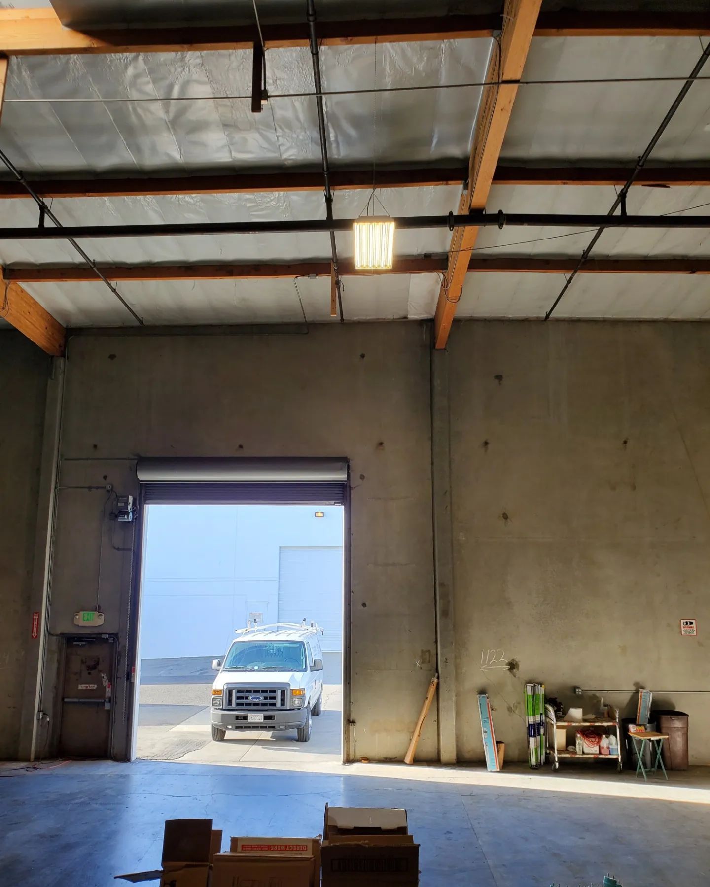Warehouse interior; an open door reveals a white van. Building materials and equipment are present.
