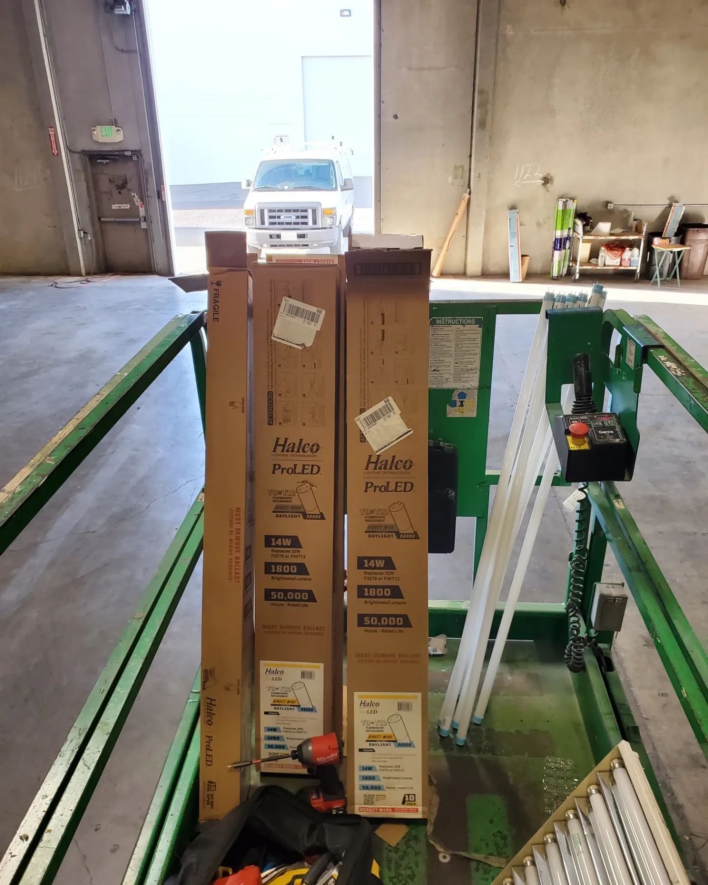 Boxes and tools on a green lift in a warehouse, with a van outside.