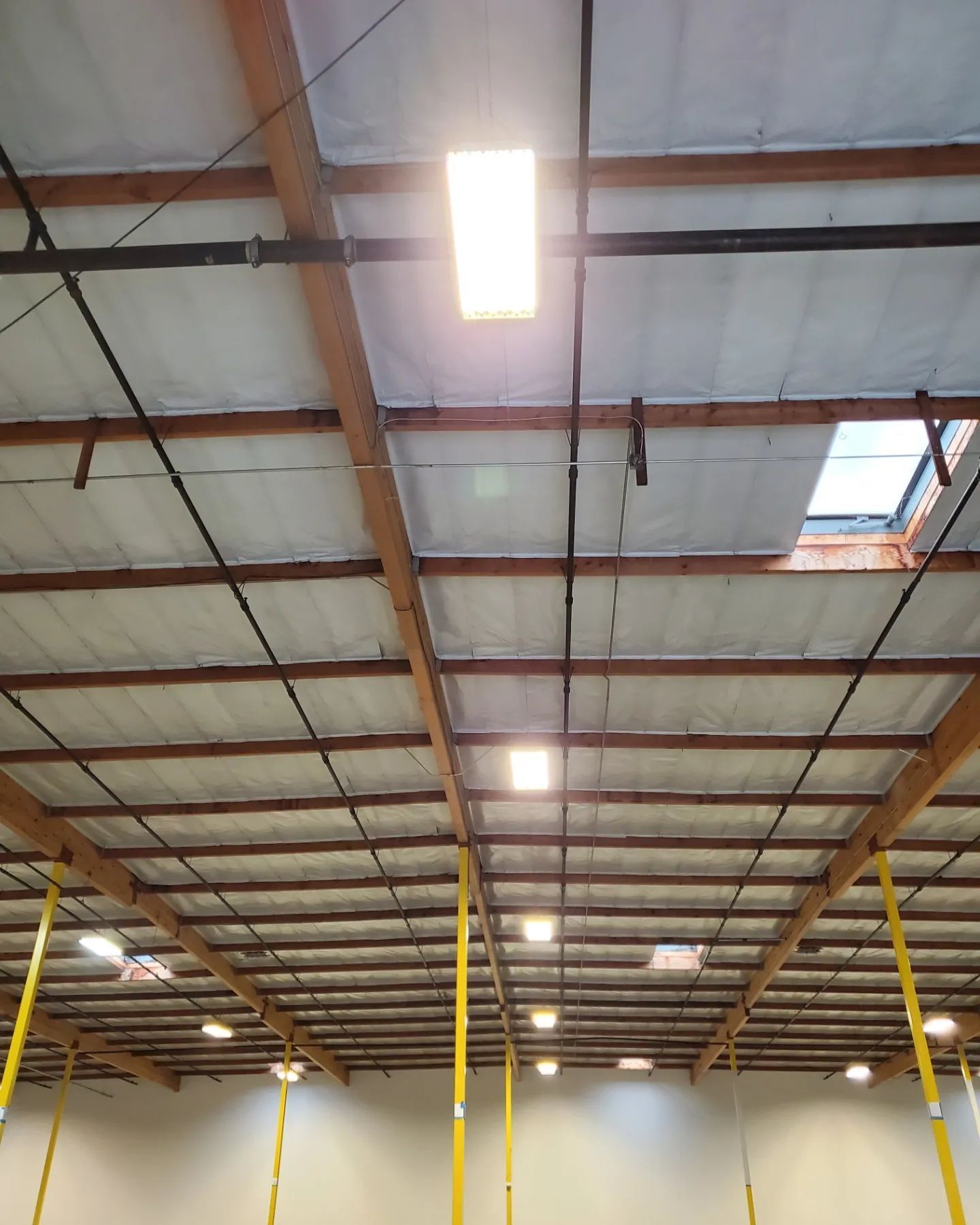 Warehouse ceiling with exposed wooden beams, bright lights, and insulation.