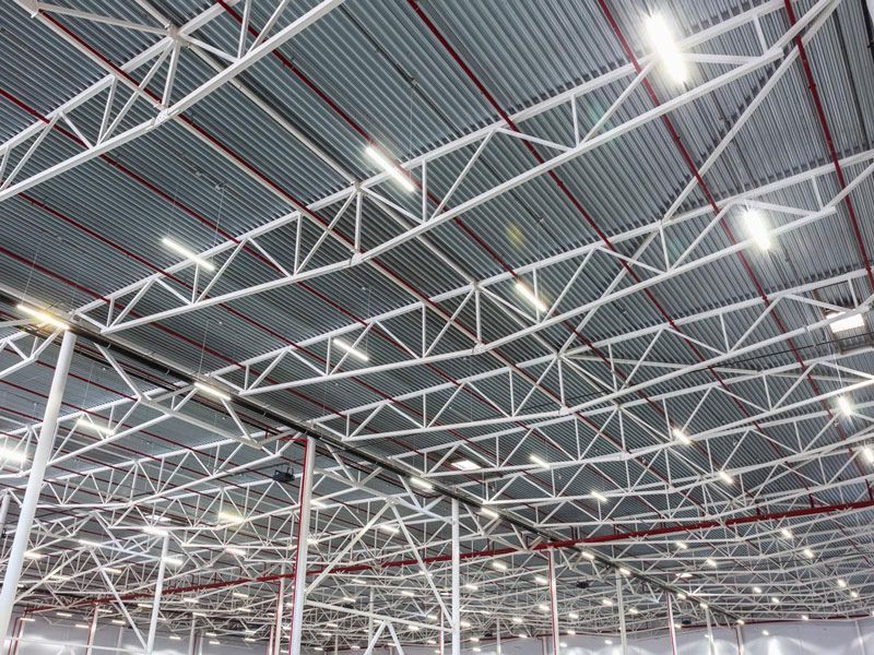 Warehouse interior with metal truss roof, corrugated metal ceiling, and overhead lighting.