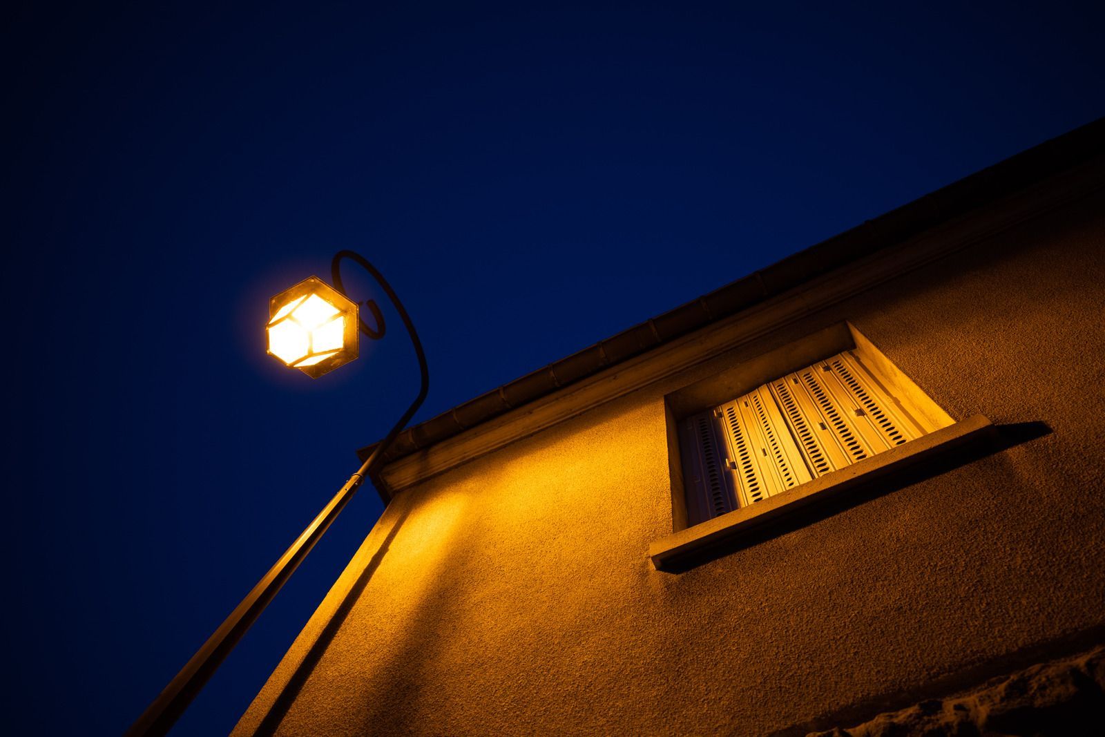A lamppost illuminates a building with a window, against a dark blue evening sky.