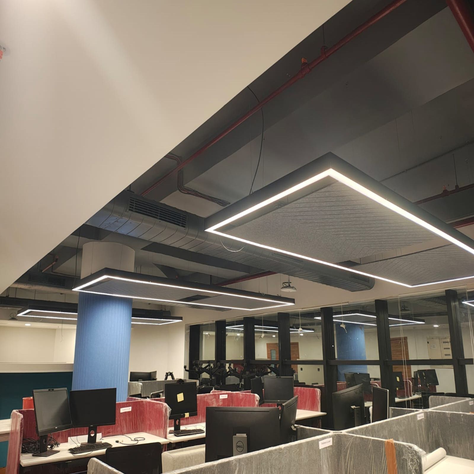 Open-plan office with desks, computers, and overhead lights. Blue column, dark ceiling, and glass walls are visible.