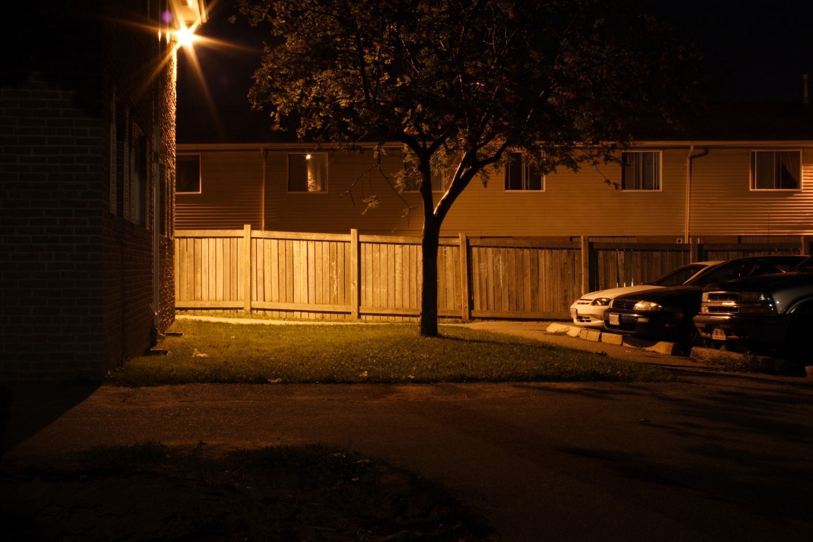 Apartment complex at night, lit by streetlight. Cars parked, fence, and tree in the foreground.