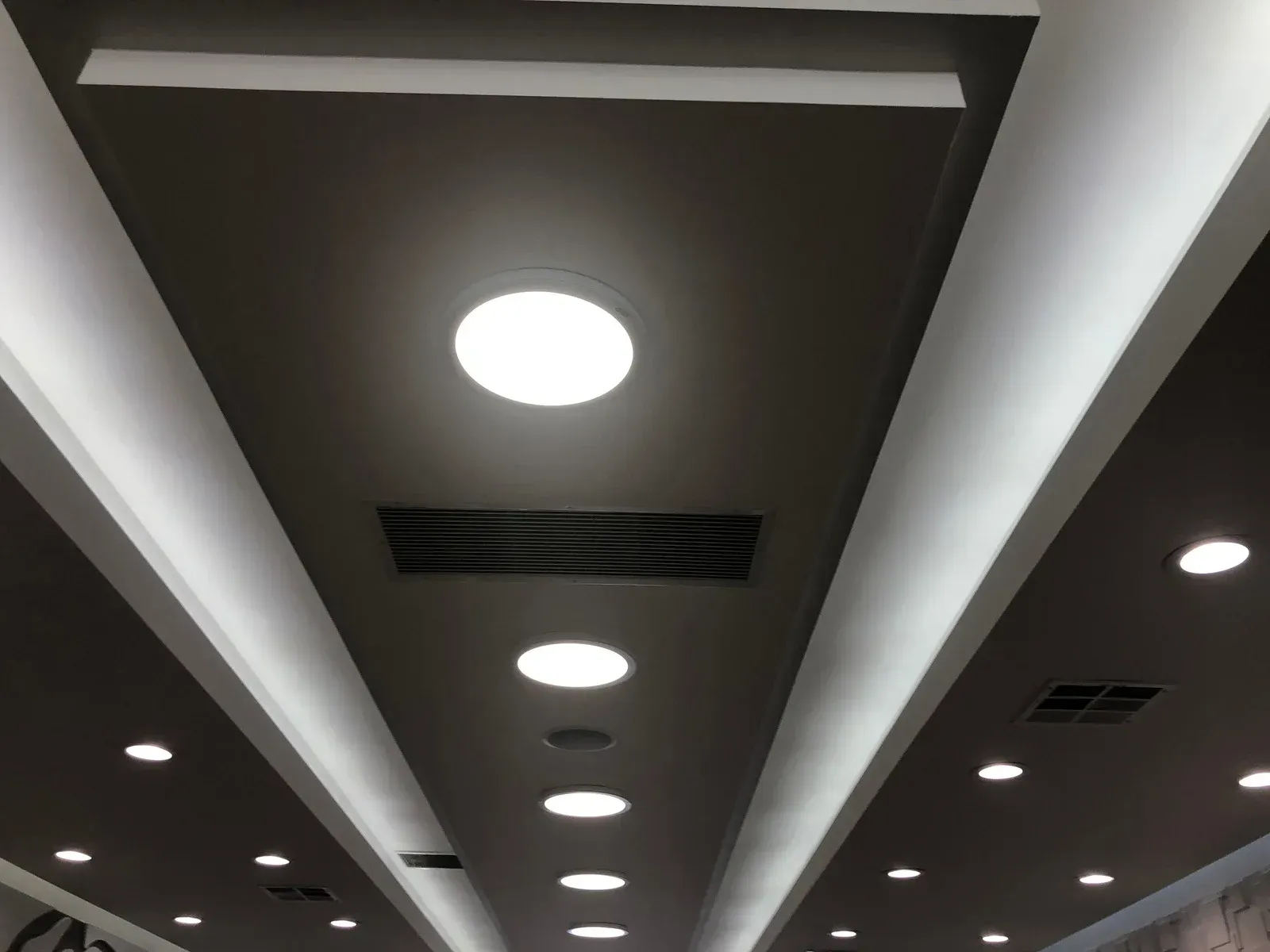 Low angle shot of a ceiling with recessed lights, ventilation grill, and white trim against a dark gray background.