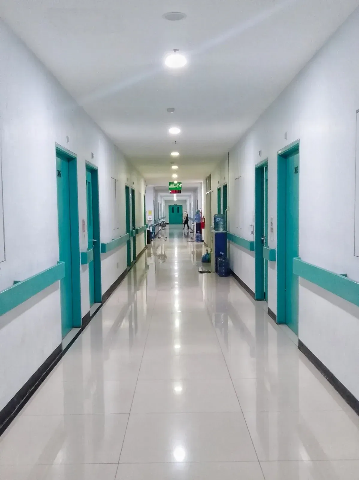 Hospital hallway with teal doors and handrails. Fluorescent lights and reflective flooring.