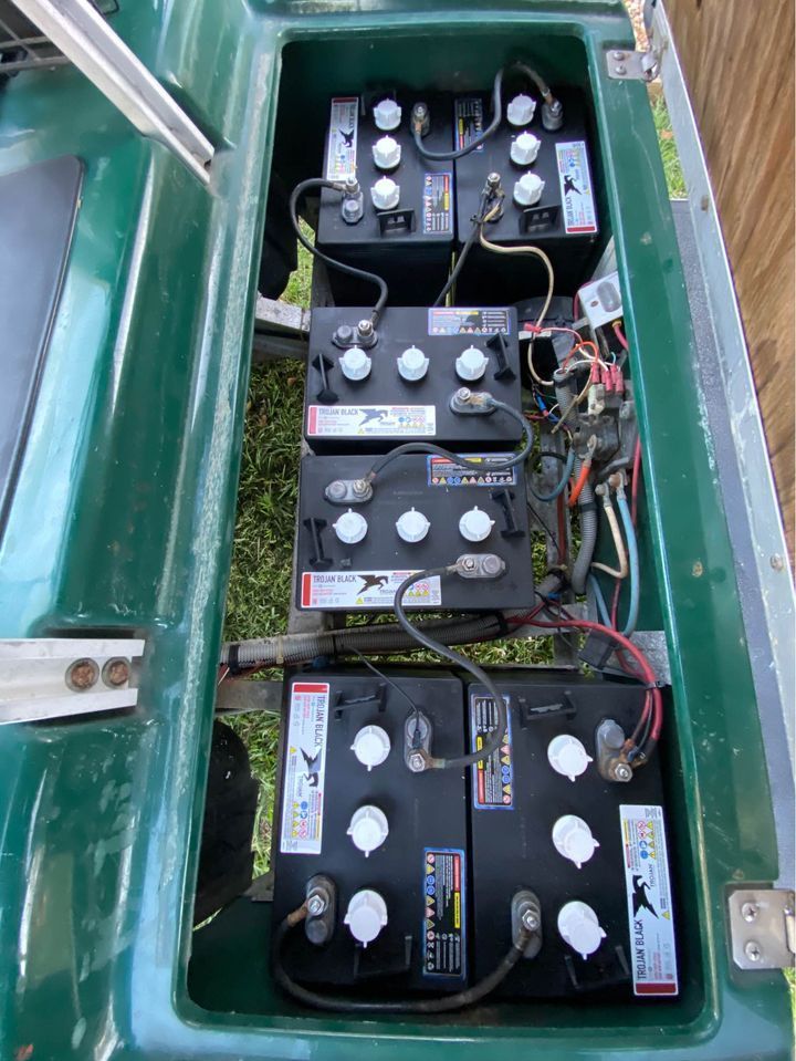 Eight black golf cart batteries, arranged in two rows, inside a green compartment. Cables connect batteries.