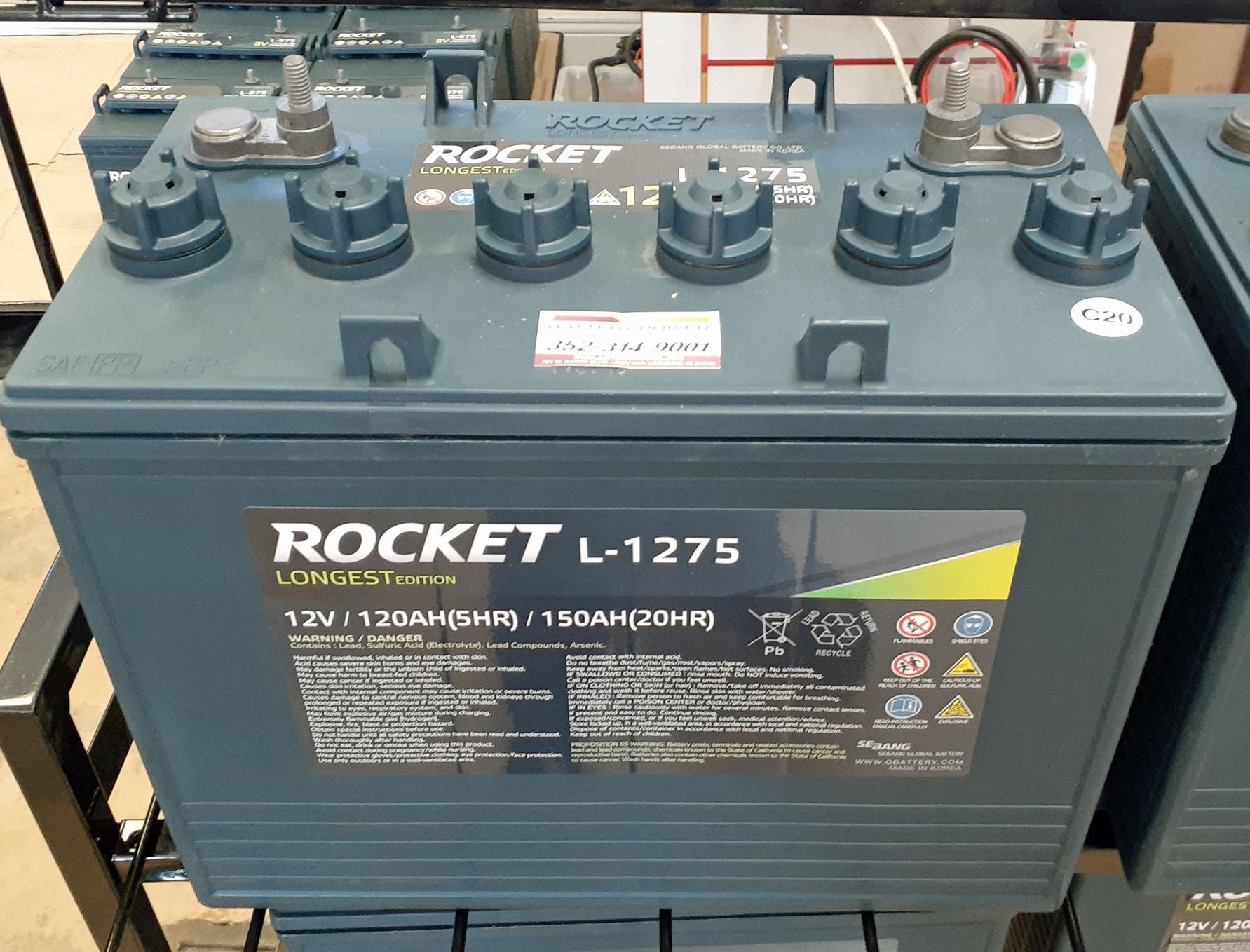 A blue Rocket L-1275 12V battery with six terminals, displayed on a shelf.