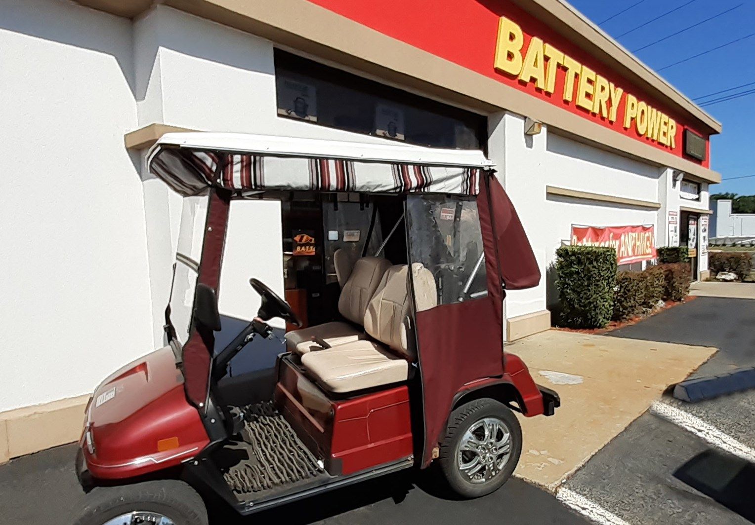 Red golf cart parked in front of a Battery Power store with a canopy and clear side panels.
