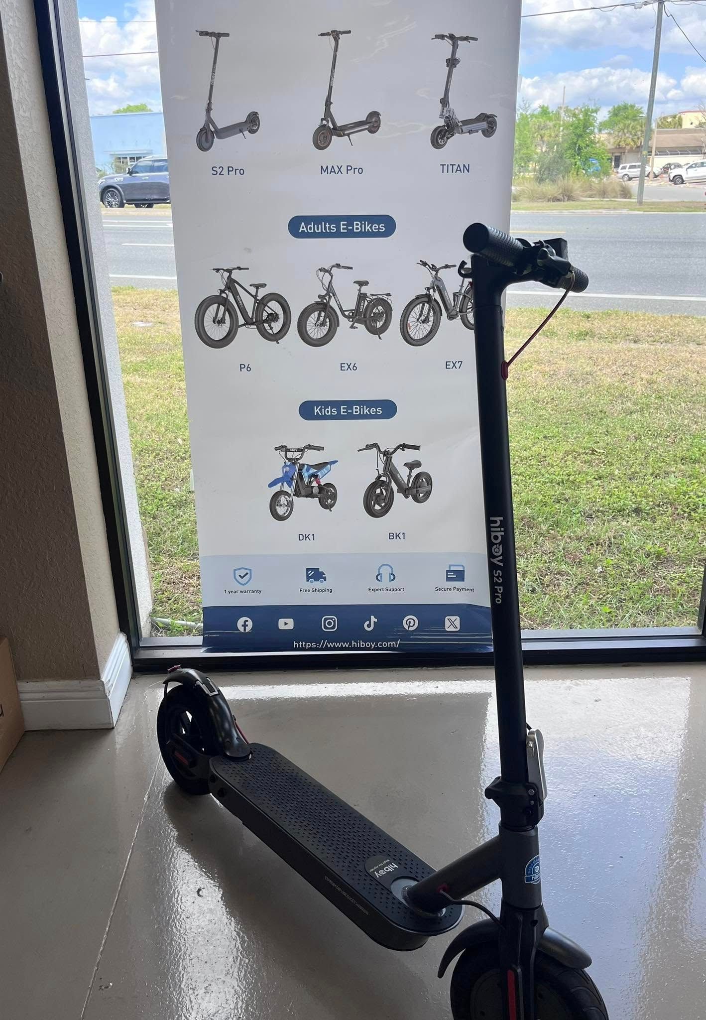 Black electric scooter in front of a banner displaying other models, near a window.