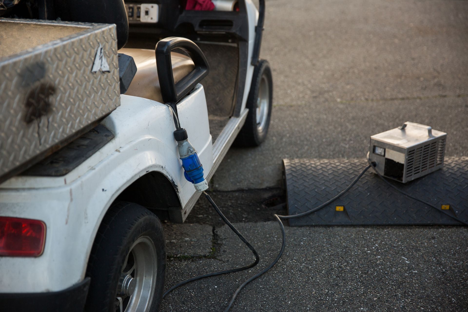 White golf cart plugged into a power source on asphalt. A metal toolbox sits on the back.