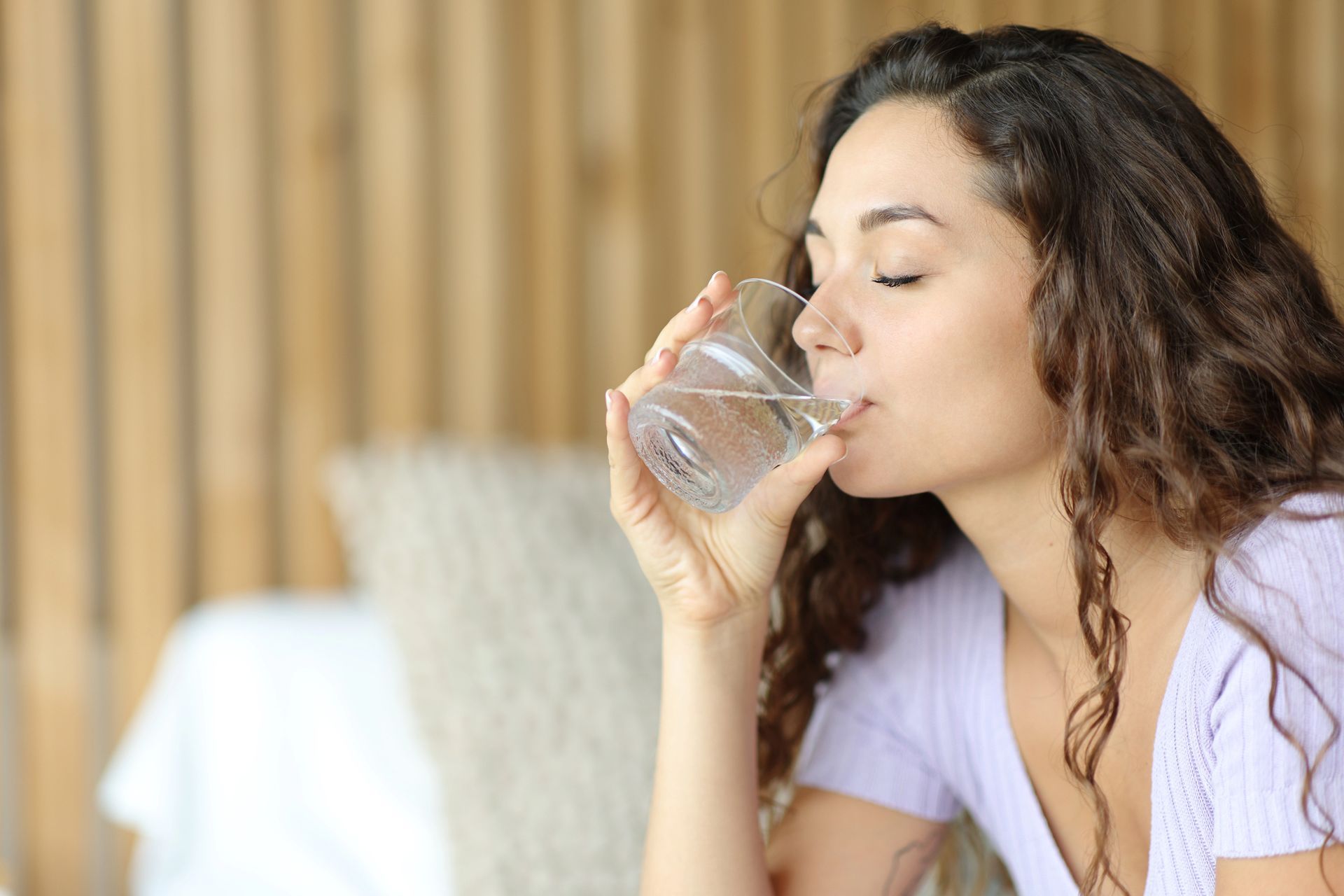 A woman is drinking a glass of water while sitting on a bed.