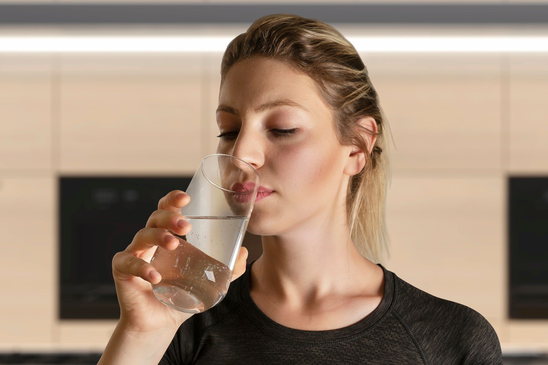 A woman is drinking a glass of water in a kitchen.