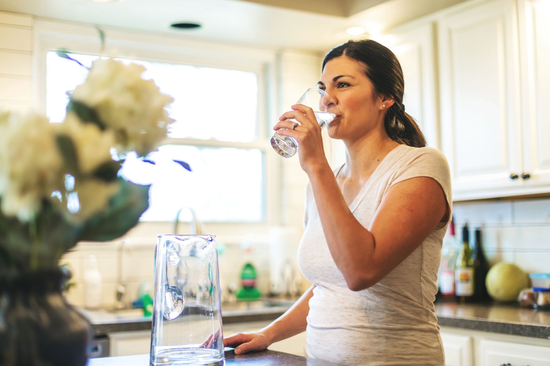 A woman is drinking a glass of water in a kitchen.