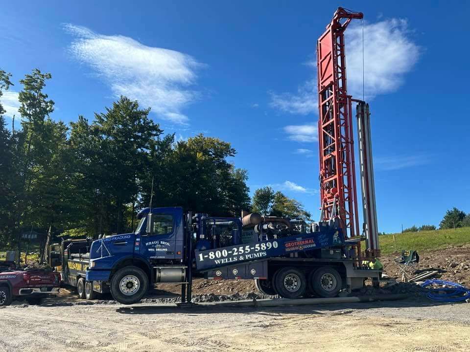 A blue well drilling truck is parked in a dirt lot with trees in the background.