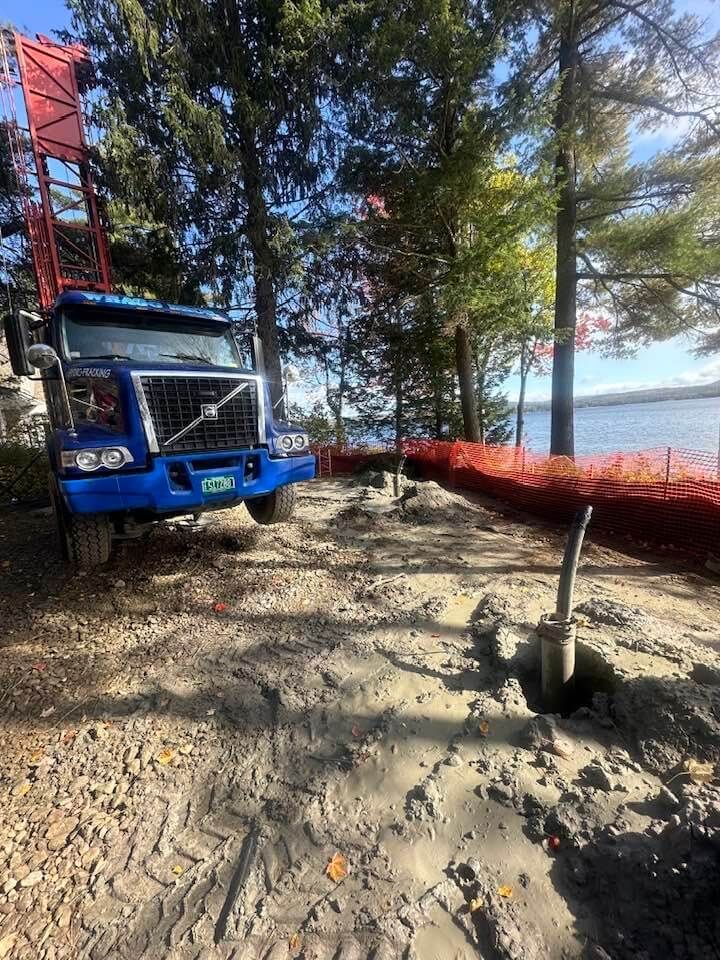 A blue well drilling truck is parked on a dirt road next to a body of water.