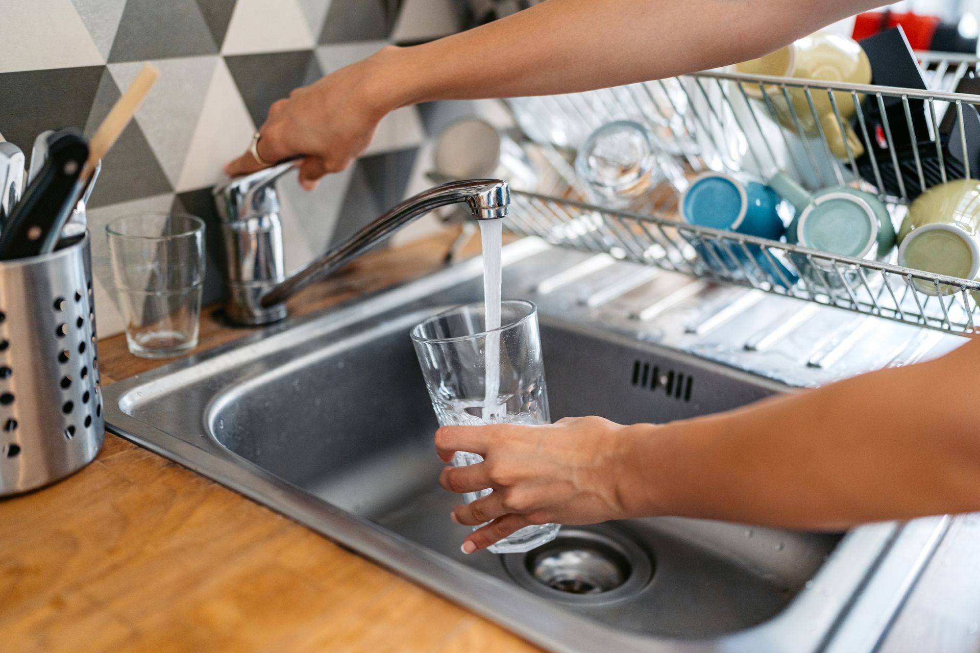 Person filling a glass with water from a kitchen faucet in a stainless steel sink.