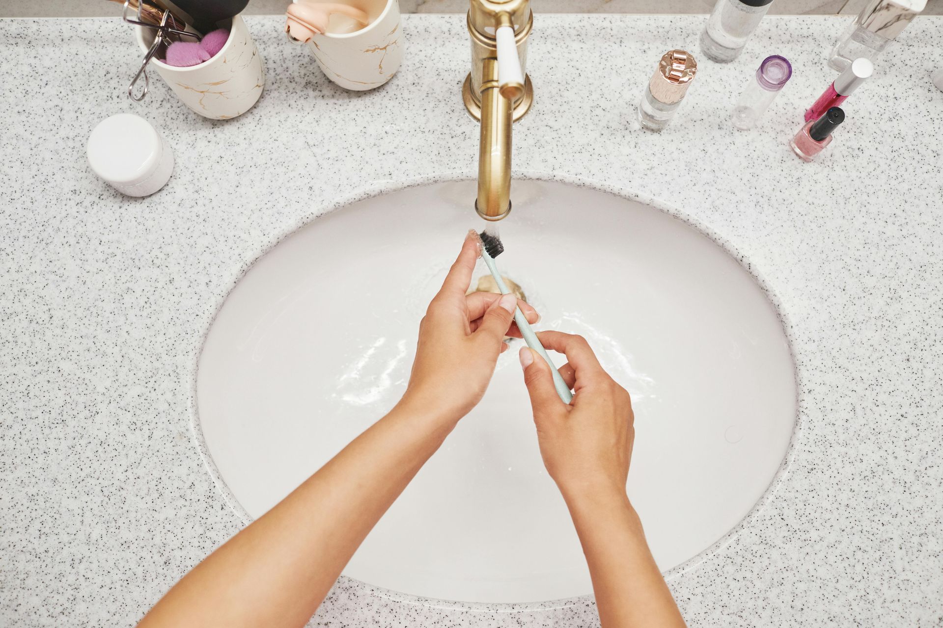 Hands rinsing a toothbrush under a gold faucet in a white bathroom sink.