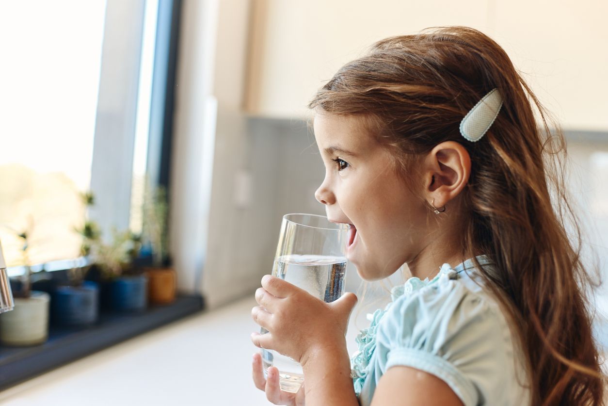 A little girl is drinking a glass of water in a kitchen.