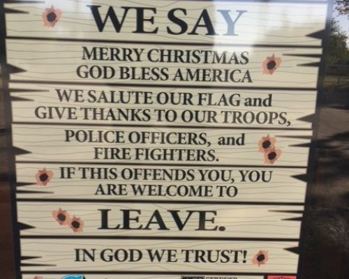 Wooden sign with patriotic and religious text, decorated with bullet holes on a weathered background.