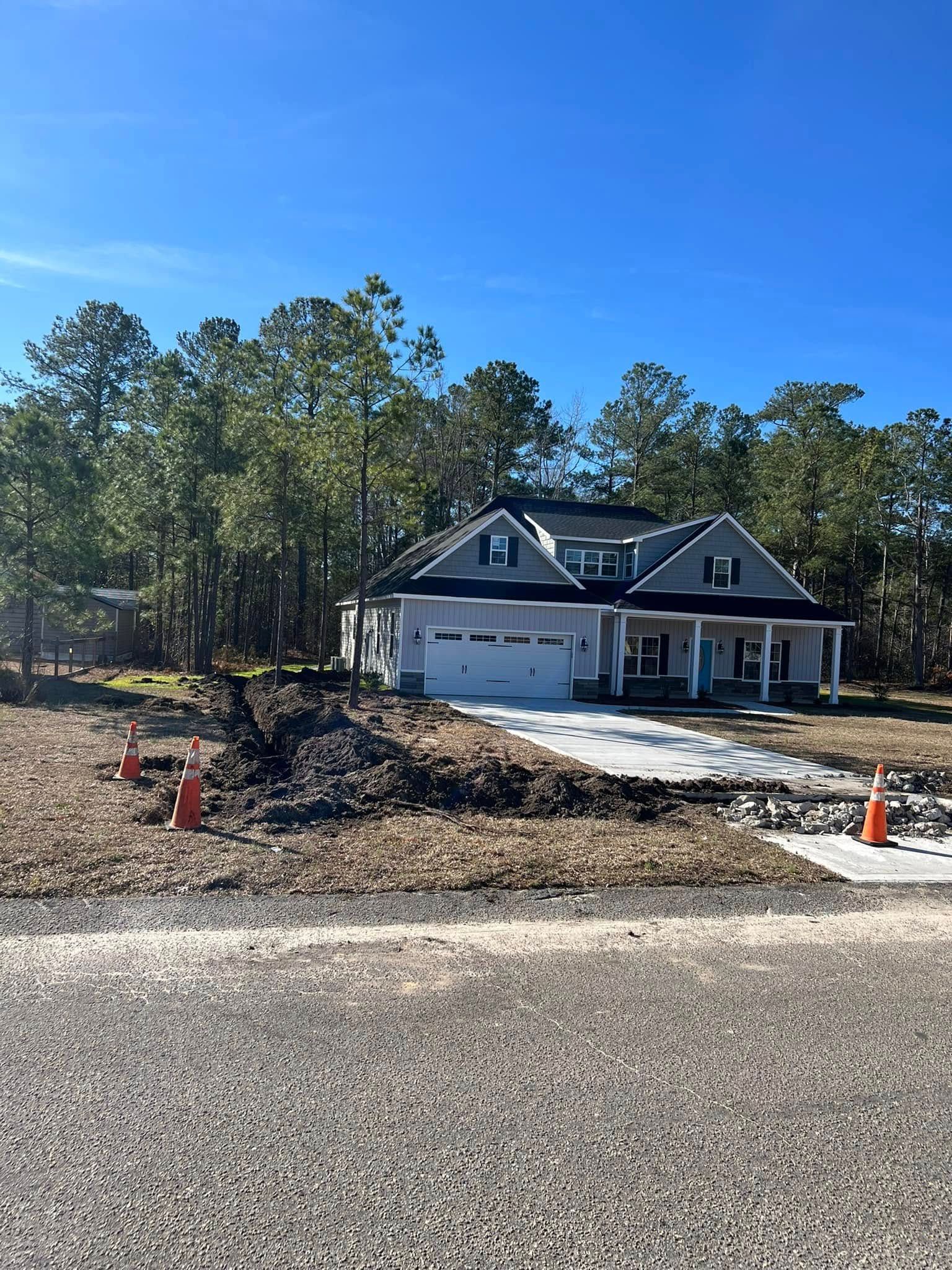 House under construction with gravel driveway, pine trees, and orange traffic cones beside a quiet road
