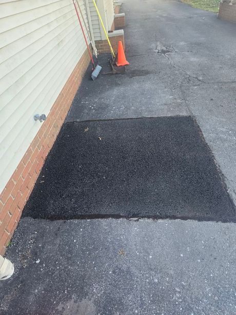 Black mulch patch on asphalt driveway next to a building. A traffic cone and broom are visible.
